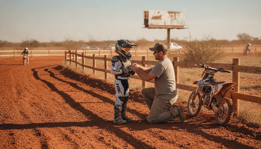 Parent helping a young motocross rider adjust protective gear beside a dirt track